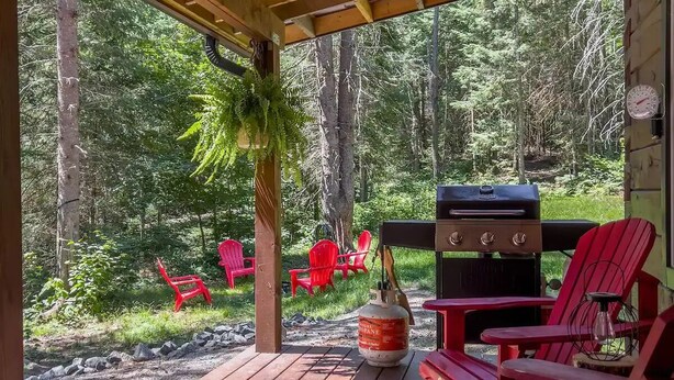 Un espace extérieur abrité doté d’une terrasse en bois, de chaises Adirondack rouges, d’un barbecue et d’une bouteille de propane.
