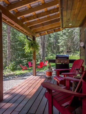 A covered outdoor area with a wooden deck, red Adirondack chairs, a grill, and a propane tank. - Charming 4 season log cabin near Algonquin Park (Kearney)