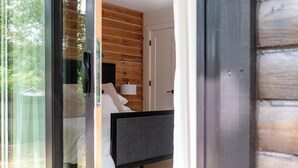 Un dormitorio moderno con puerta de cristal que da a un balcón, una cama con ropa de cama blanca, una mesita de noche con lámpara y paneles de madera en las paredes.