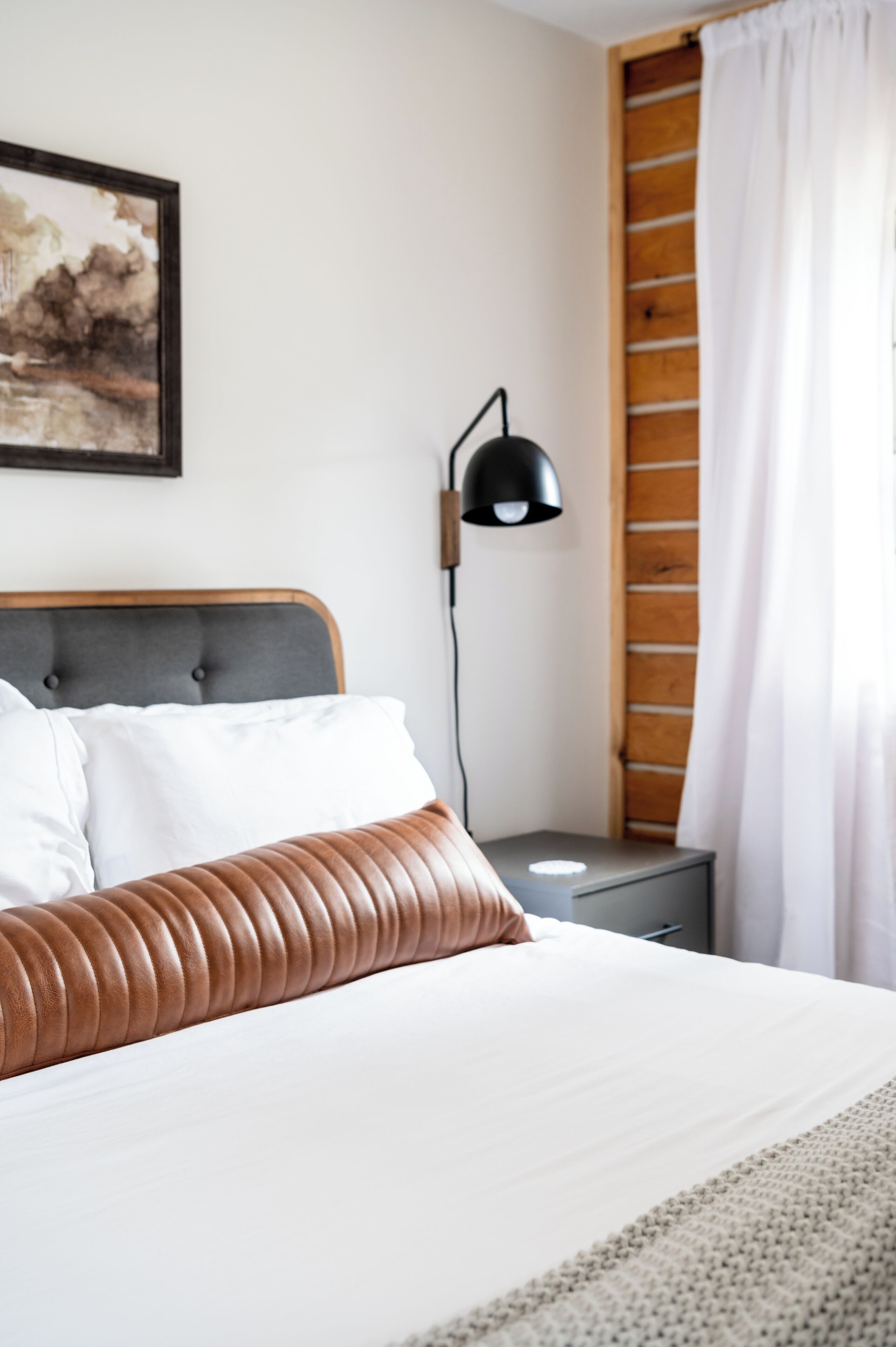 Cama bem arrumada com cabeceira de madeira e detalhes em couro marrom, mesa de cabeceira com abajur e um quadro na parede.