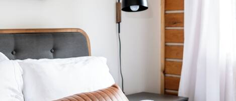 Cama bem arrumada com cabeceira de madeira e detalhes em couro marrom, mesa de cabeceira com abajur e um quadro na parede.