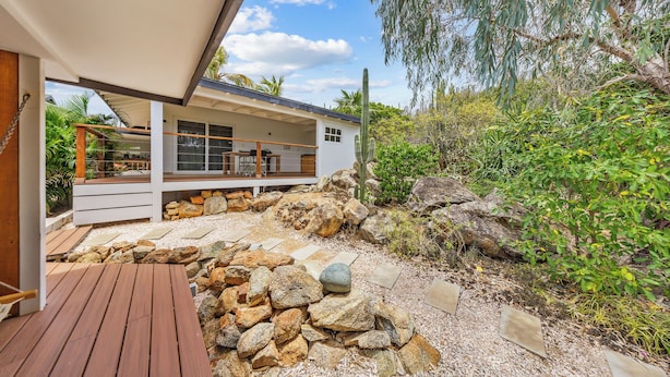 A modern house with a wooden deck, a stone pathway, and a variety of plants and trees.