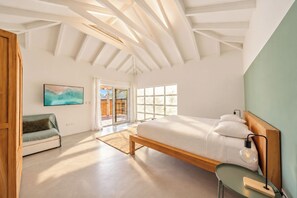A bedroom with a large bed, a wooden headboard, a desk with a lamp, a sofa, and a sliding glass door leading to a balcony.