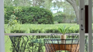 Una mesa de vidrio con una silla de mimbre, una planta en maceta y vistas a un jardín cuidado con setos y árboles.