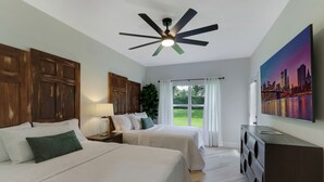 Habitación de hotel con dos camas, ventilador de techo, televisión y un amplio ventanal con cortinas.