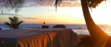 A beach with a bed under a palm tree, overlooking the ocean at sunset.