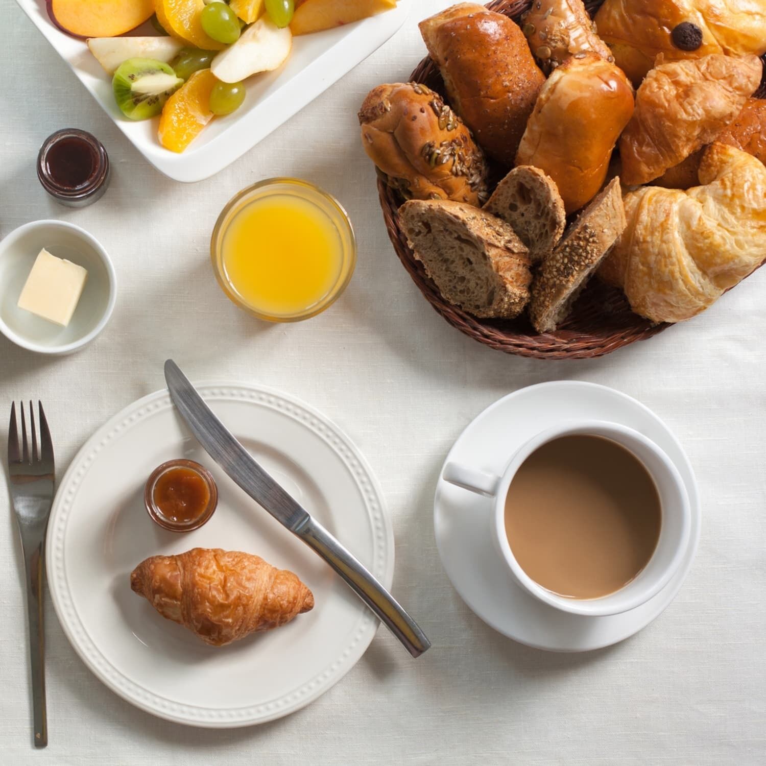 Aamiaispöytä, jossa on croissantteja, leipää, hedelmiä ja kuppi kahvia valkoisella pöydällä.