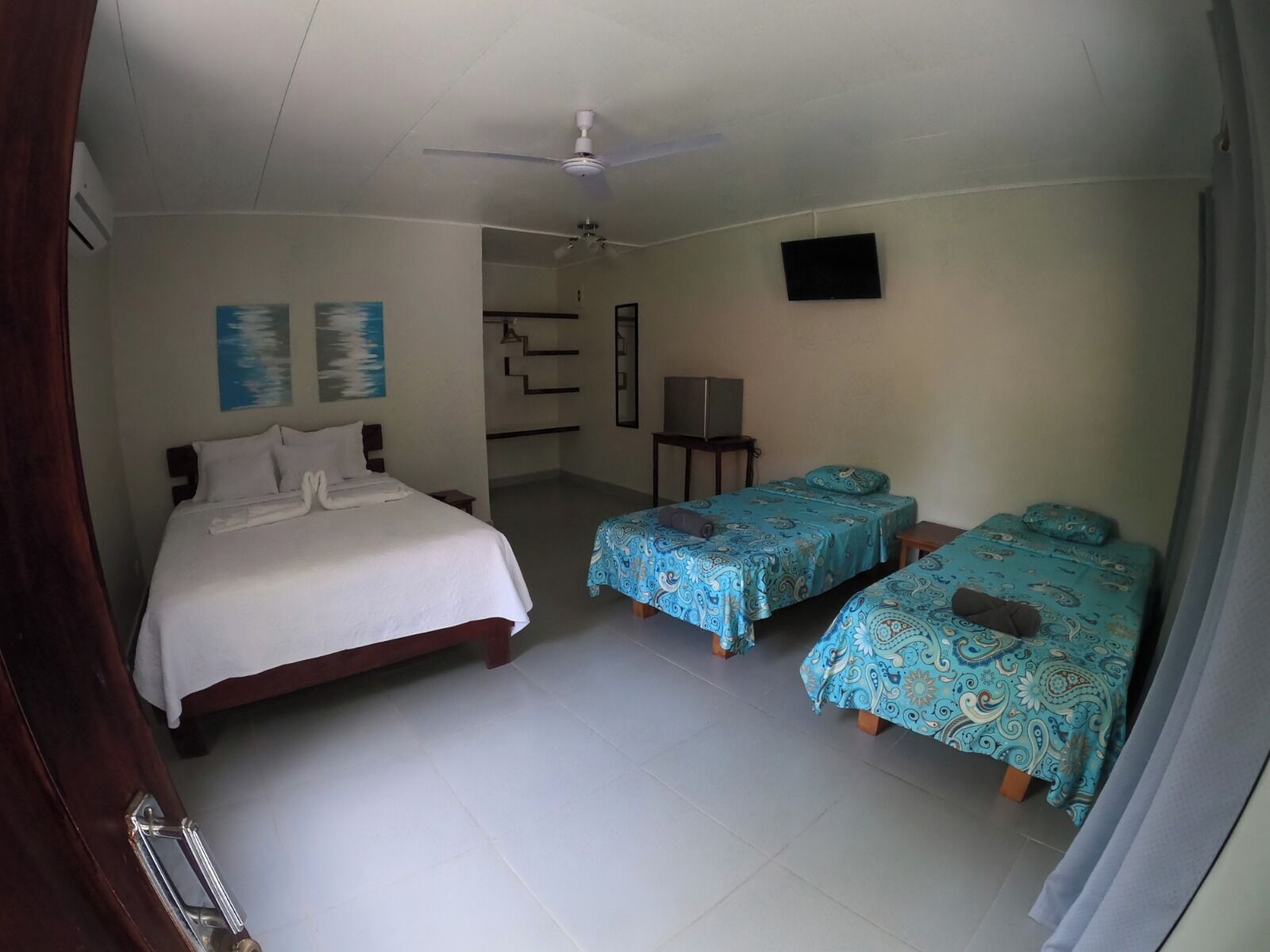 A hotel room with a large bed, two smaller beds, a ceiling fan, a TV, and artwork on the walls.