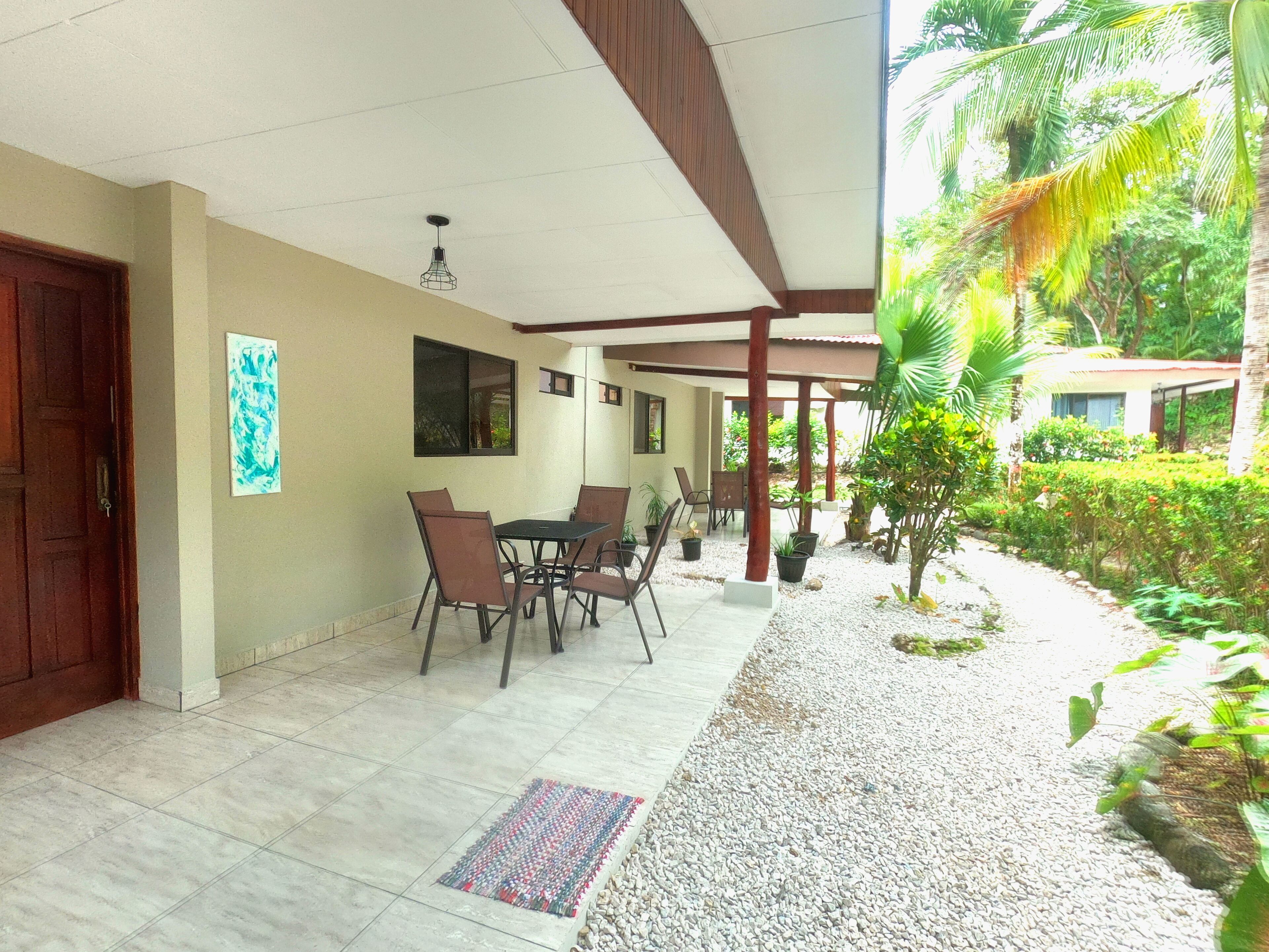 A covered patio with a tiled floor, a dining table with chairs, and a small garden area with plants and a palm tree.