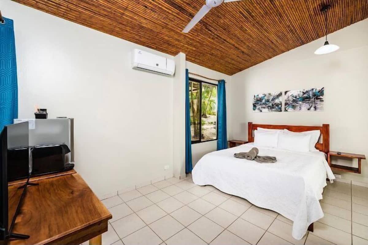 Een slaapkamer met een bed, een televisie op een houten tafel, een plafondventilator en een raam met blauwe gordijnen.