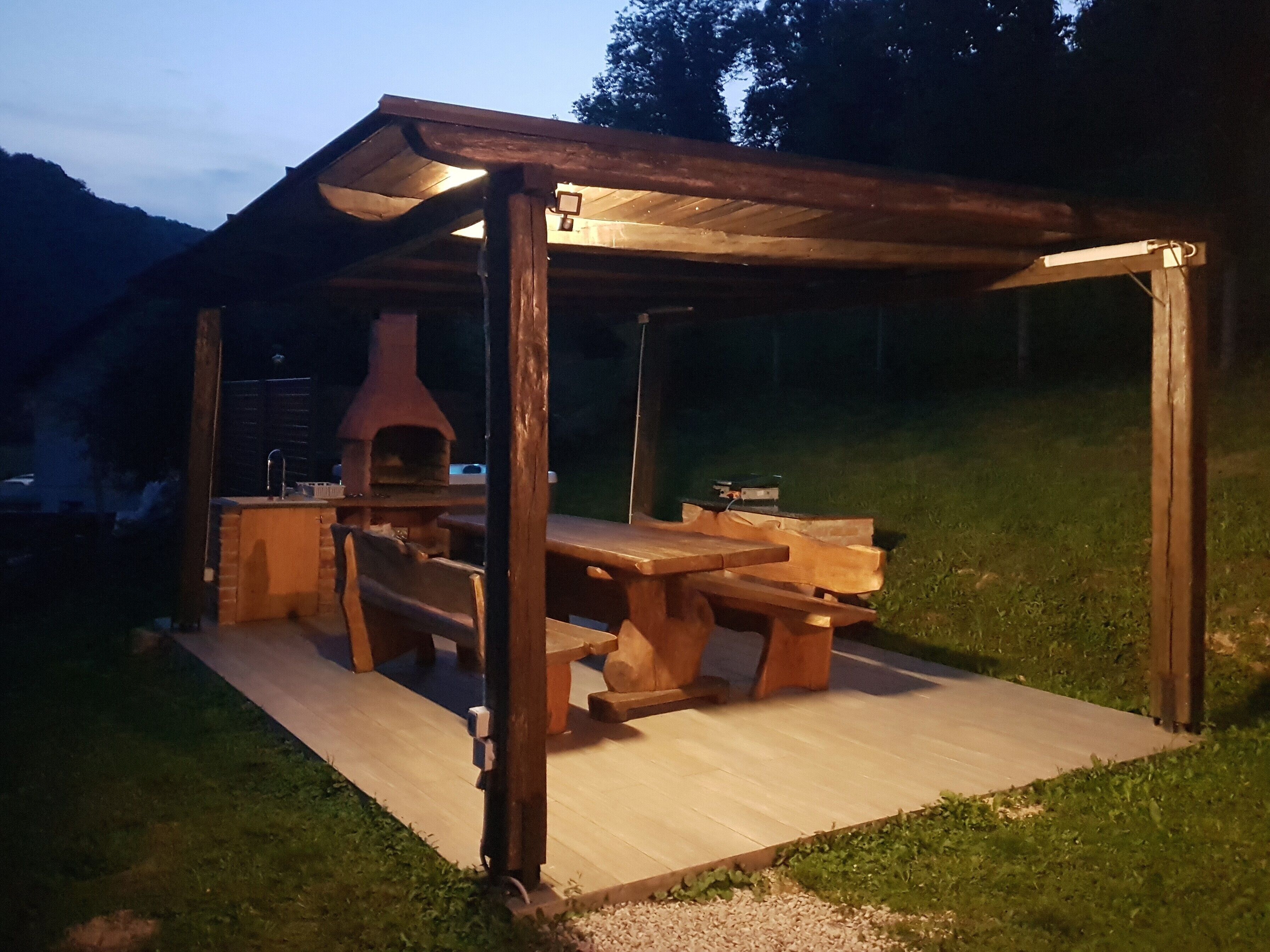 Een houten constructie buiten, met een ingebouwde pizzaoven, picknicktafel en banken.