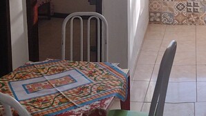 Un comedor con una mesa cubierta por un mantel colorido, una silla con cojín verde y una silla de metal. La habitación tiene baldosas estampadas en las paredes y un suelo de baldosas.