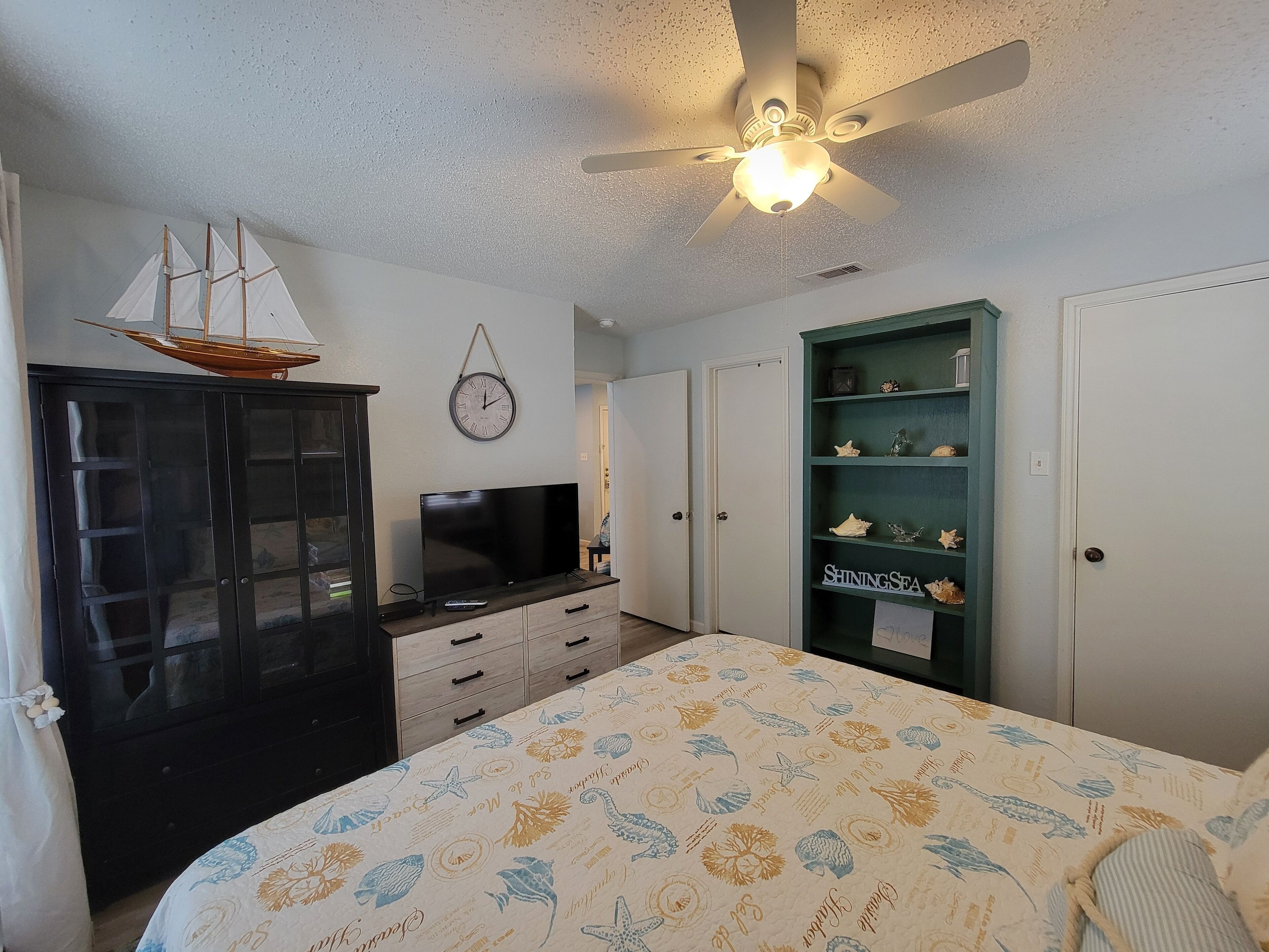 A bedroom with a bed, a TV, a wardrobe, a ceiling fan, and a shelf with decorative items.