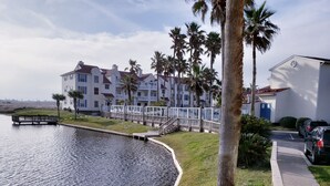 Une propriété située au bord de l’eau, dotée d’une terrasse en bois, d’une allée pavée et d’une rangée de bâtiments blancs avec des balcons.