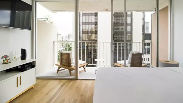 Una habitación de hotel moderna con una cama grande, un televisor, un balcón con una silla y una mesa, y vistas a la ciudad.