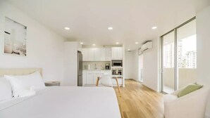 Una habitación de hotel moderna con una cama grande, una mesa de comedor, una zona de cocina y vistas a la ciudad a través de amplios ventanales.