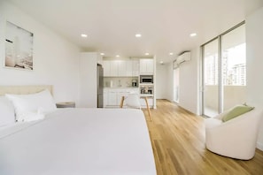 A modern hotel room with a large bed, a dining table, a kitchen area, and a view of the city through large windows. - Modern Design Studio in Waikiki w/ FREE PARKING (Honolulu)