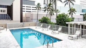Una piscina en la azotea con agua azul cristalina, rodeada de suelo de baldosas blancas y barandillas metálicas. Se observan varias sillas de descanso y una zona de estar cubierta.