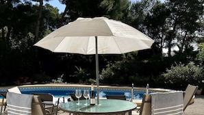 Eine Terrasse im Freien mit einem runden Tisch und Stühlen, einem großen Sonnenschirm und einem Pool im Hintergrund.
