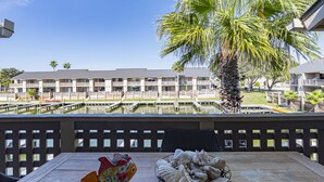Eine Holzterrasse mit einer verzierten Fischfigur und einem Korb voll Meeresschalen, mit Blick auf einen Pool und ein Gebäude mit Balkonen.