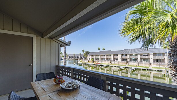 Une table en bois, entourée de chaises, est placée sur un balcon qui donne sur une marina où sont amarrés des bateaux, avec un bâtiment visible au loin.