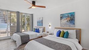 Habitación de hotel con dos camas, ventilador de techo, obras de arte en las paredes y vistas a la playa a través de amplias ventanas.