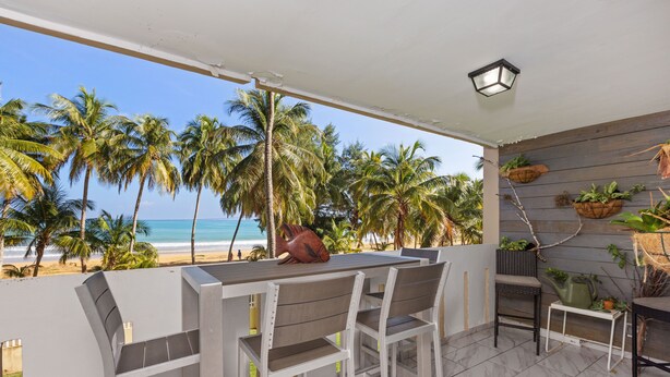 Un patio al aire libre con vista a la playa, que incluye una mesa y sillas, una mesita con una planta, y una jardinera de pared con follaje.