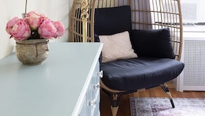 A room with a wicker chair, a desk with drawers, a vase with flowers, a lamp, and a window with a curtain. - Classy 2BR/1,5BA Boutique at Flushing! (Queens)