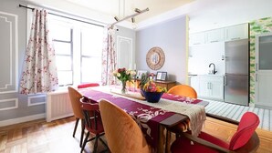 A dining area with a table set for four, surrounded by red chairs, a bowl of fruit, and a vase of flowers. A kitchen with white cabinets and a refrigerator is visible in the background. - Classy 2BR/1,5BA Boutique at Flushing! (Queens)