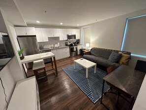 A modern kitchen with white cabinets, a refrigerator, and a microwave. There is a dining area with a white table and a gray sofa. - Newly furnished 1 bed, steps to Boston's restaurants, museums, hospitals, Fenway (Boston)