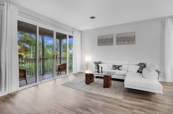 A spacious living room with a white sofa, a coffee table, and a view through large sliding glass doors to a balcony with a chair.