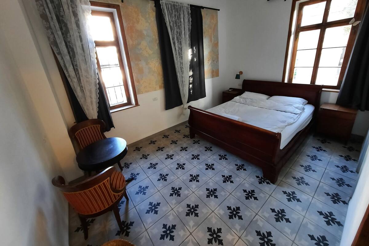 A bedroom with a wooden bed, a small table, two windows with curtains, and patterned floor tiles.