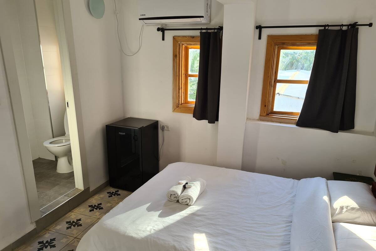 A small, clean room with a bed, a black refrigerator, a toilet, and a window with curtains.