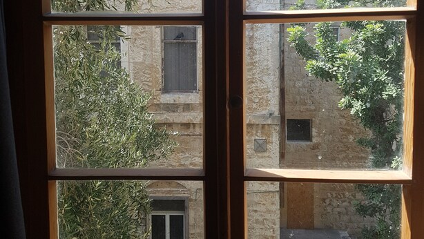 A window with multiple panes, each offering a view of a building with stone walls and greenery. - Haifa Vintage Rooms (Haifa)