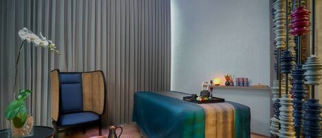 A room with a bed, a chair, a small table with a plant, and a decorative wall piece.
