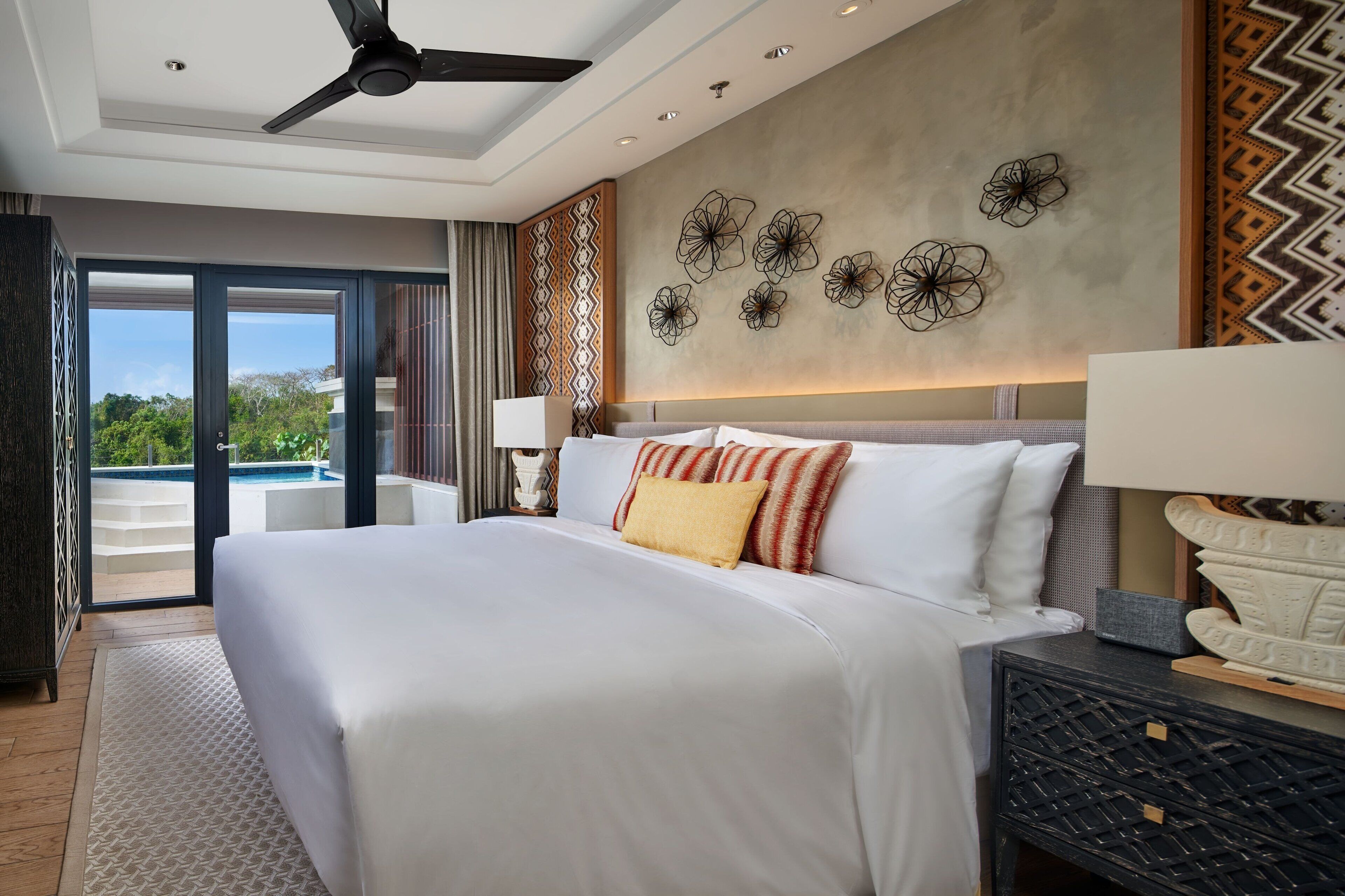 Habitación de hotel con una cama grande, ventilador de techo, vista al exterior y pared decorativa con motivos florales.