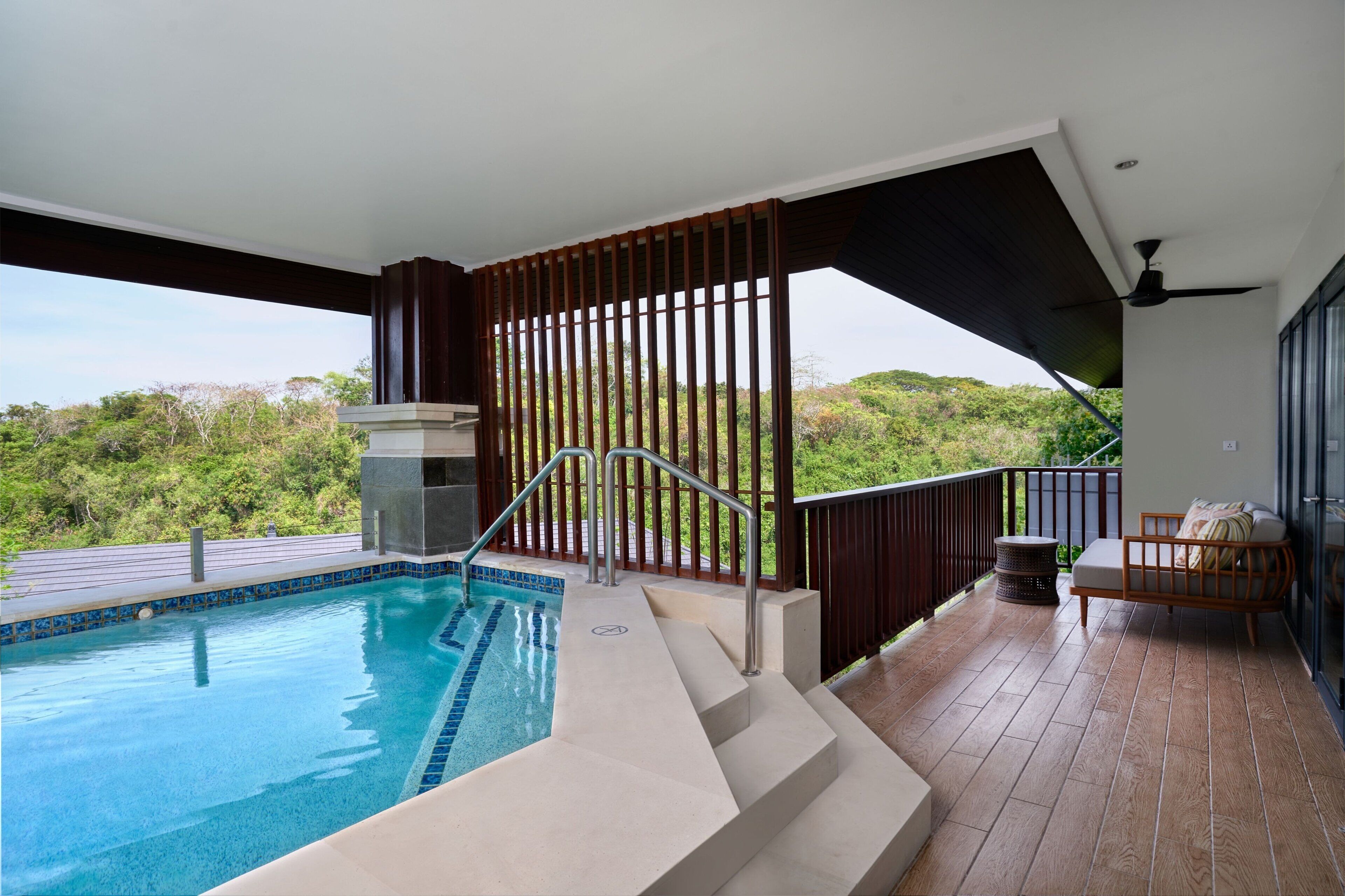 Zona de piscina con escalones que llevan al agua, una terraza de madera y un área de descanso con vistas a la vegetación.