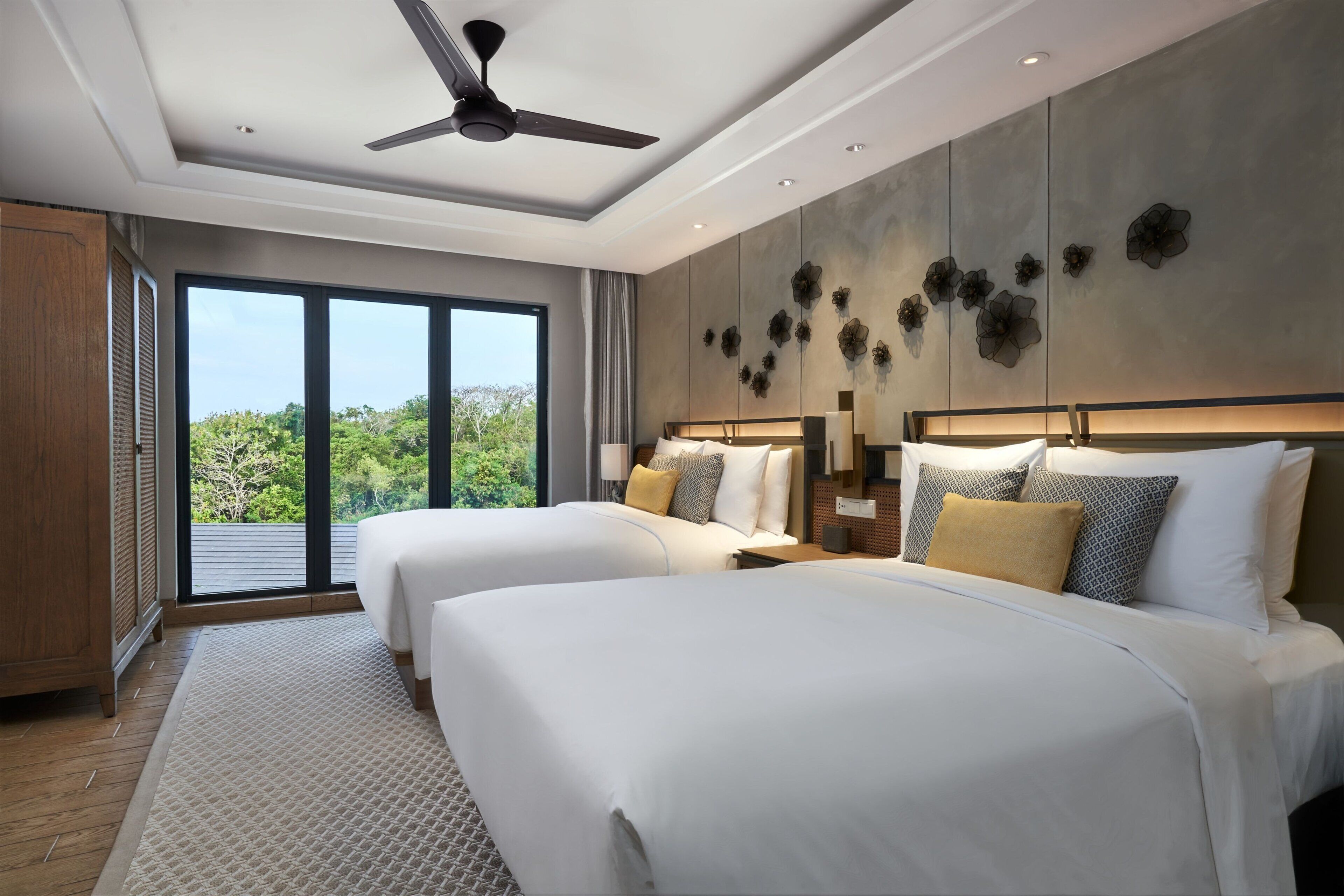 A modern hotel room with two beds, a ceiling fan, large windows, and a view of greenery.