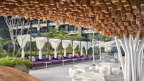 An outdoor seating area with white tables and chairs, a wooden ceiling structure, and a building in the background. - Marriott's Bali Nusa Dua Terrace (Nusa Dua)