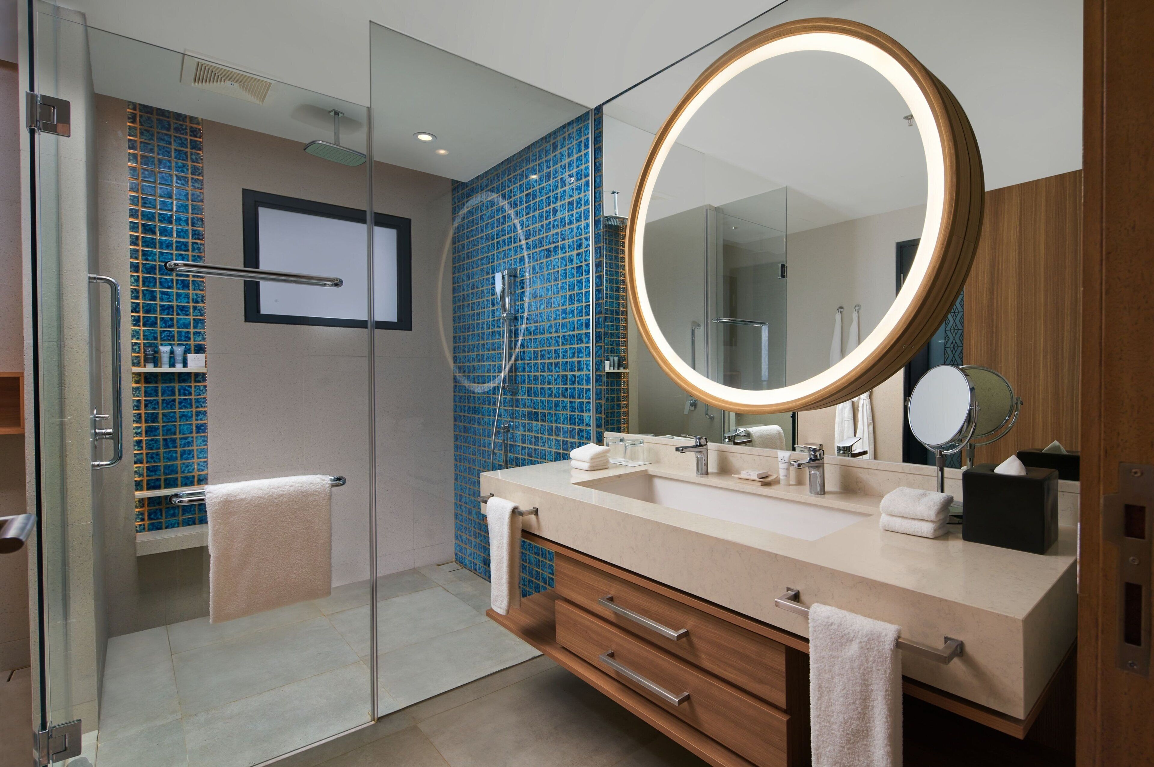 A modern bathroom with a glass shower enclosure, a large round mirror, and a beige vanity with a towel rack.