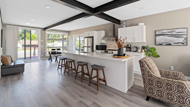 A modern kitchen with white cabinets, a central island with stools, and a dining area with a gray sofa.