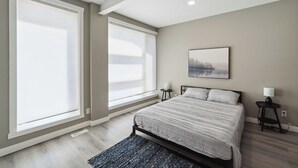 A bedroom with a bed, two bedside tables, a nightstand, a window with blinds, and a painting on the wall.