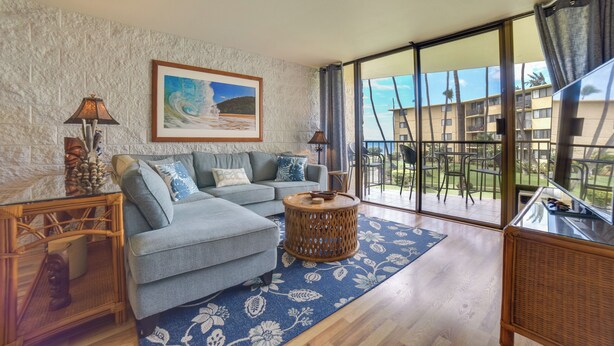 A living room with a grey sofa, a round wooden side table, and a large window with a view of a building and greenery. - Maalaea Condo w/ Pool Access - Walk to Beach! (Wailuku)