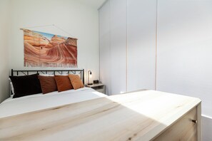 A bedroom with a bed, a nightstand, a lamp, and a large painting of a canyon. - Modern Studio with Kitchen, Washer/Dryer, Fire Pit, Grill, & Red Rock Views! (Kanab)