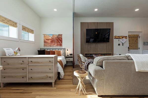 A modern bedroom with a bed, a sofa, a wooden dresser, and a television mounted on the wall. - Modern Studio with Kitchen, Washer/Dryer, Fire Pit, Grill, & Red Rock Views! (Kanab)