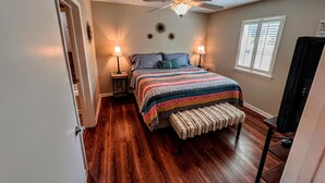 A bedroom with a bed, two bedside lamps, a ceiling fan, a window with blinds, and a television on a stand.