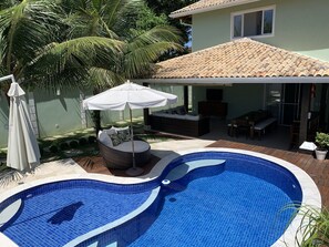 Piscina com borda azulejada, área de estar coberta com mesa e cadeiras, e um grande guarda-sol.