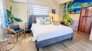 A bedroom with a bed, a ceiling fan, a window with blinds, a wall mural, and a door. - Costa Nube (Caye Caulker)