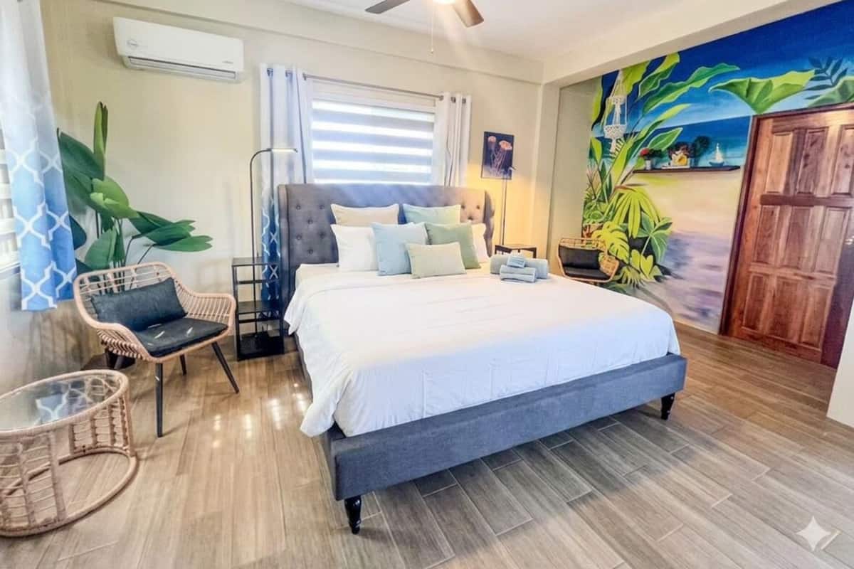 A bedroom with a bed, a ceiling fan, a window with blinds, a wall mural, and a door.