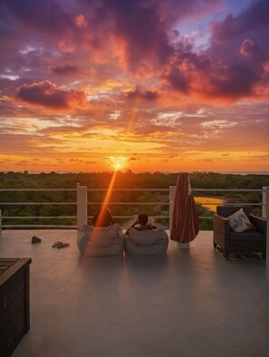 Vista al atardecer desde un balcón con puffs, una silla y un tejido colgante.